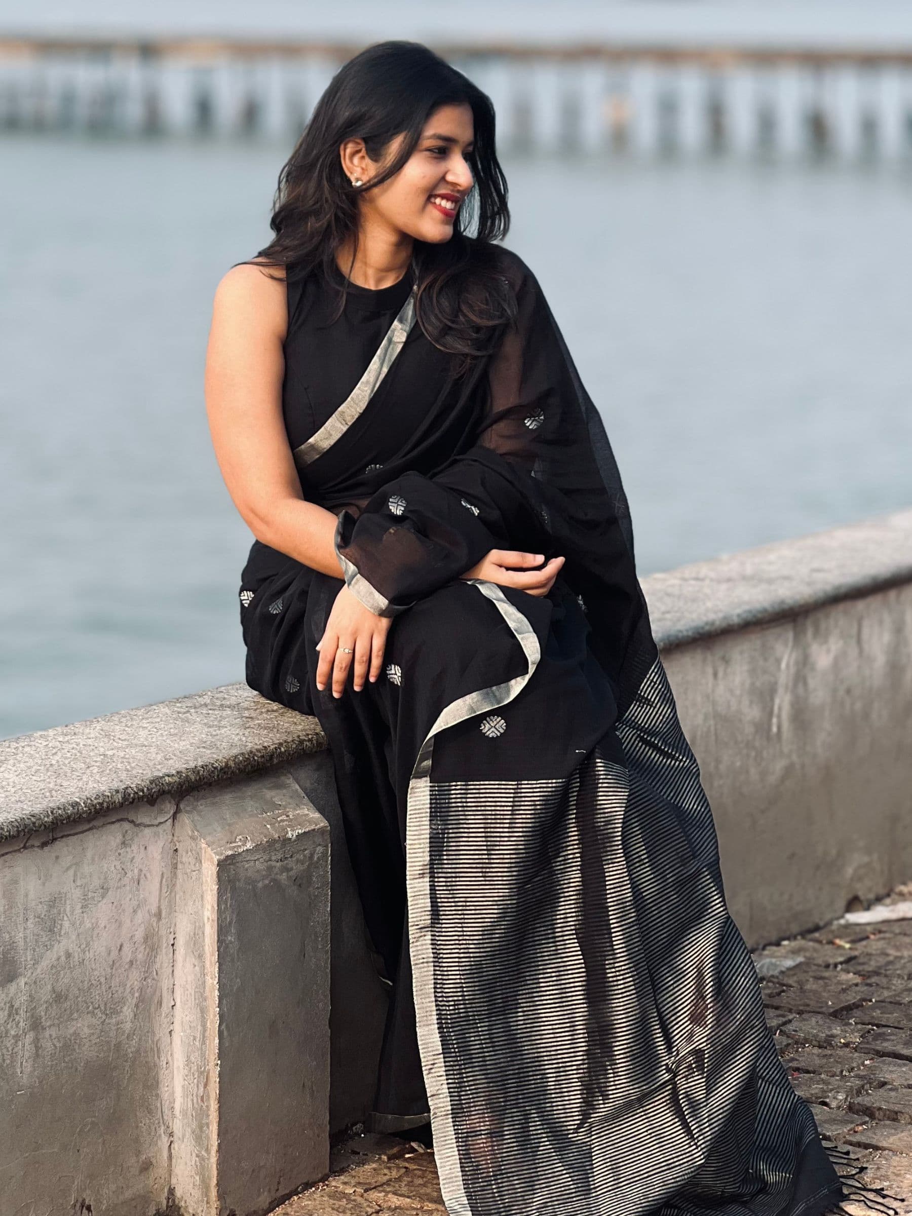 Woman in a black saree with silver border sitting by a waterfront.